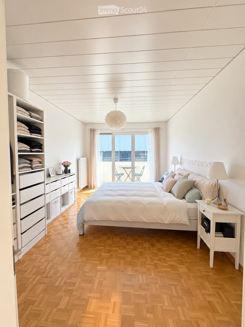 Bright, modern bedroom with wooden herringbone flooring, large bed with pillows and blankets, nightstand with lamp, storage unit with folded blankets, and a small seating area with a table and chairs near the window.