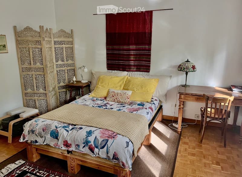 Cozy bedroom with a wooden bed, yellow pillows, floral duvet, nightstand with lamp, desk with chair, and decorative screen.