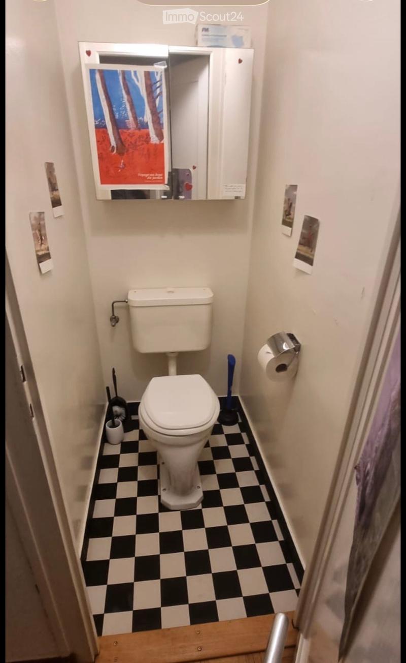 Small bathroom with white walls, black and white checkered floor, toilet, toilet paper holder, toilet brush, and a mirror with a poster on it.