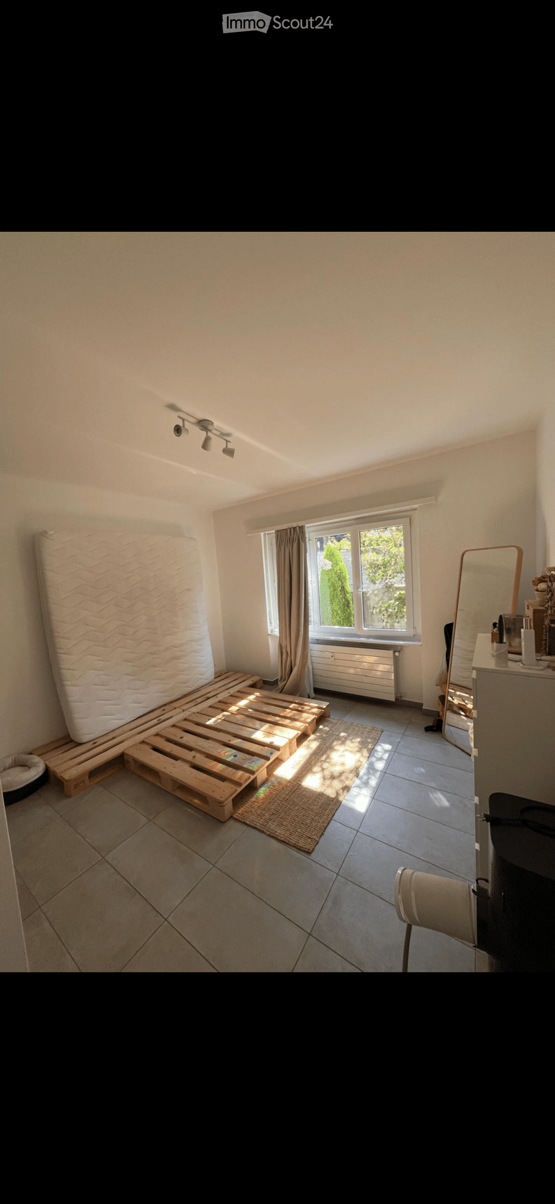 A furnished bedroom with a wooden pallet bed, a chair, a mirror, a radiator, and a window with curtains. The room has tiled flooring and a small rug.