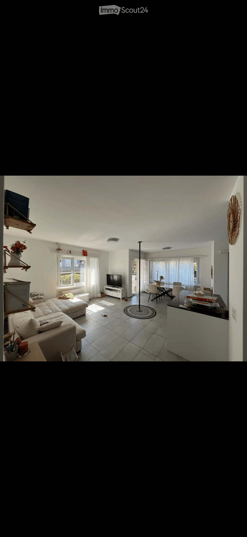 Bright living room with a large sofa, coffee table, TV stand, dining area with table and chairs, and decorative shelf with flowers.