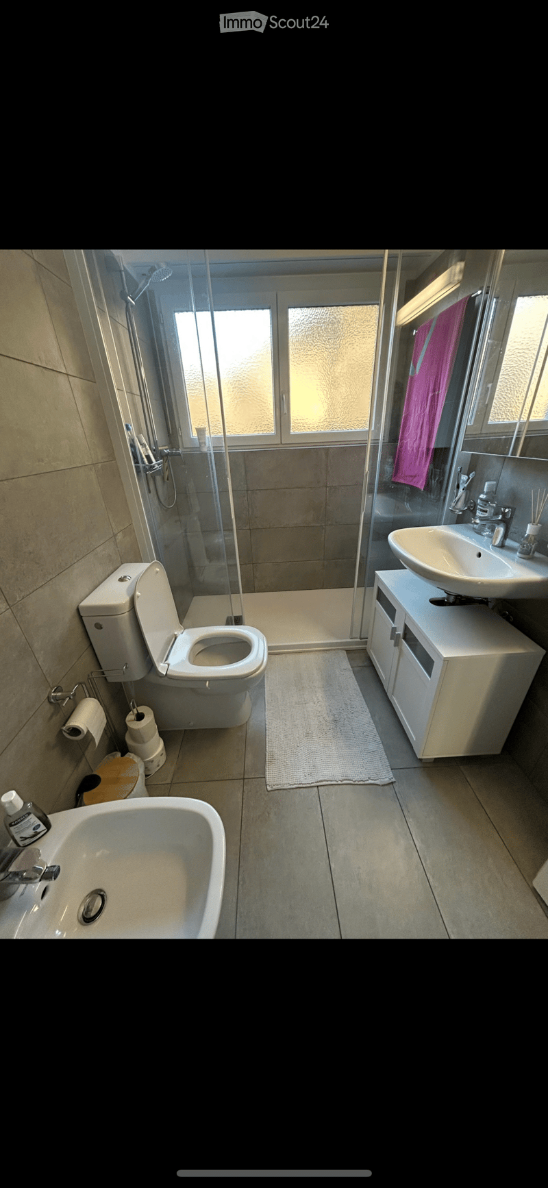 Modern bathroom with a toilet, bathtub, sink, and shower area. Features a white cabinet, mirror, and a pink towel hanging on the shower door.