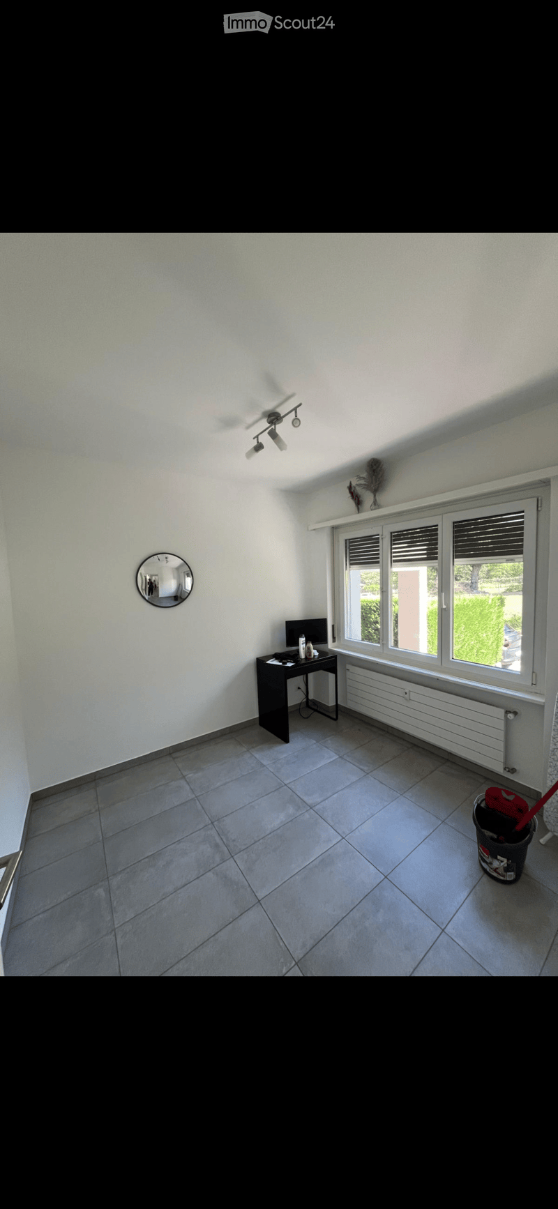 A modern, unfurnished room with a tiled floor, large windows, a desk with a computer, a mirror on the wall, and a radiator under the windows.