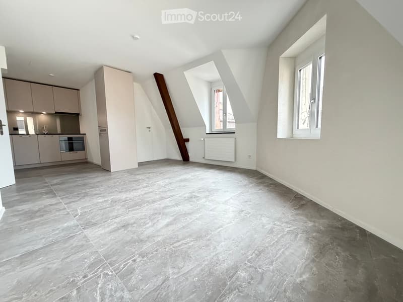 An unfurnished room with grey tiled flooring, white walls, and large windows. The room features a slanted ceiling and a small kitchen area with cabinets and appliances visible in the background.