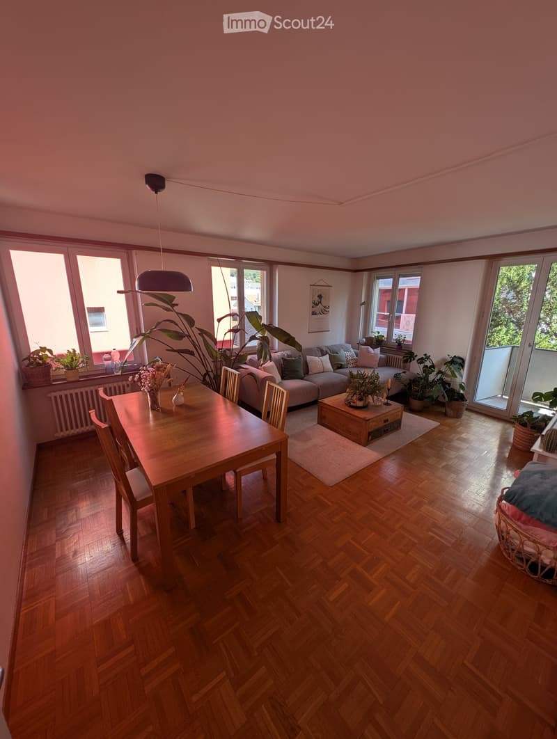 Open-plan living and dining area with wooden flooring, large windows, a sofa, dining table, chairs, coffee table, pendant light, and several plants.
