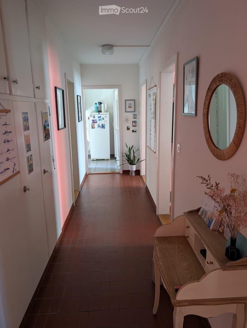 A well-lit hallway with tiled flooring, light pink walls, and several doors leading to different rooms. A small wooden bench and a console table with a vase of flowers are placed on the right side. A round mirror and framed pictures adorn the walls.