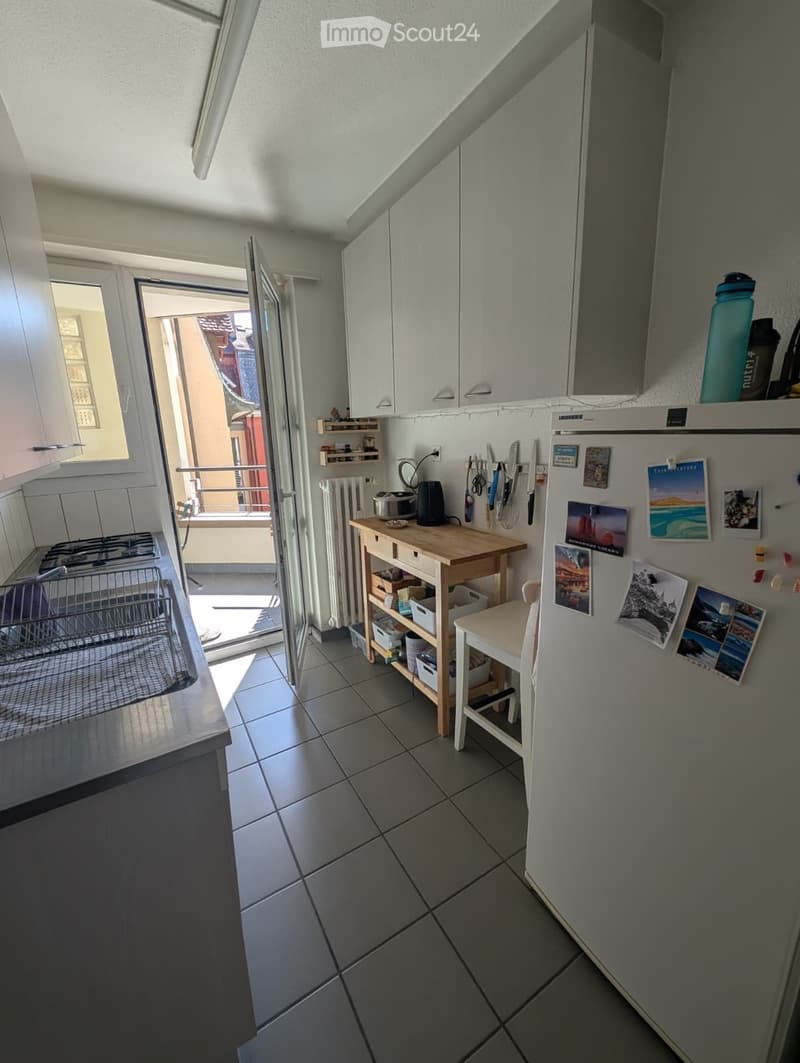 Modern kitchen with white cabinets, tiled floor, and a small wooden table with a stool. A refrigerator is adorned with various pictures and magnets. An open door leads to a balcony.