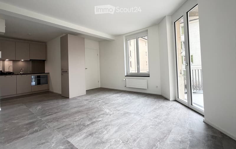 Open-plan living and kitchen area with grey tiled flooring, white walls, fitted kitchen cabinets, sink, and oven. Large window and sliding glass door leading to a balcony.