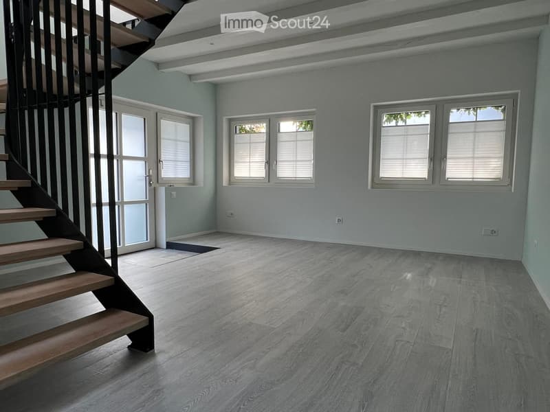 Empty room with light grey walls, wooden flooring, and multiple windows with white frames. A staircase with wooden steps and a metal railing is visible on the left side.