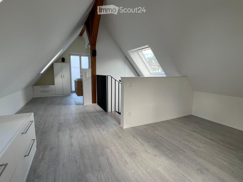 Bright, empty attic room with wooden flooring, white walls, and skylight windows. Features built-in white cabinetry and a small wooden desk area.