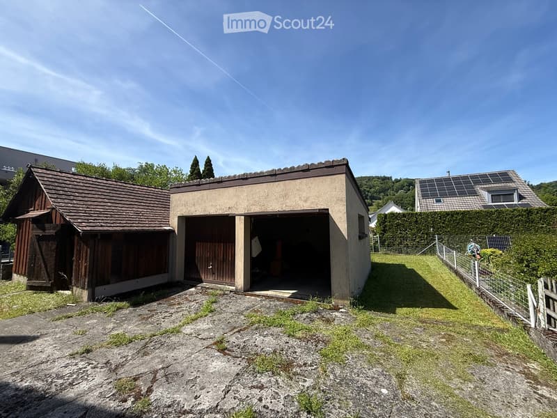 A small, single-story building with a garage and a wooden shed attached, surrounded by a grassy area and a fence.
