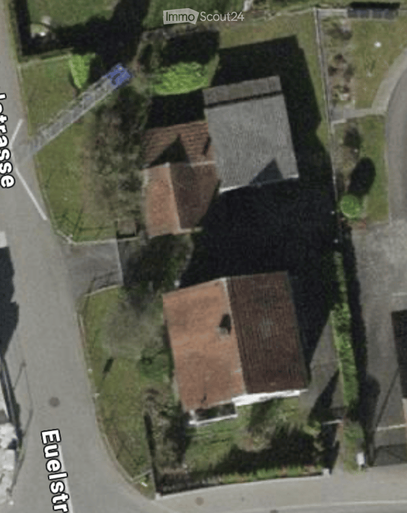 Aerial view of a residential building with a red-tiled roof, surrounded by greenery and a paved area.