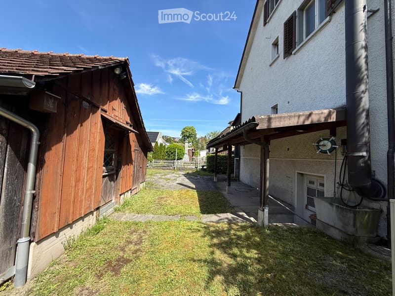 Courtyard area between two buildings, one wooden and one concrete, with grass and a clear sky.