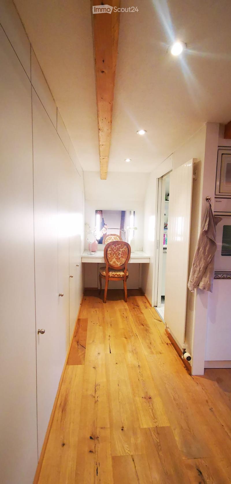 A narrow hallway with wooden flooring, white walls, and built-in cabinets on one side. A small vanity area with a chair and mirror is visible at the end of the hallway.