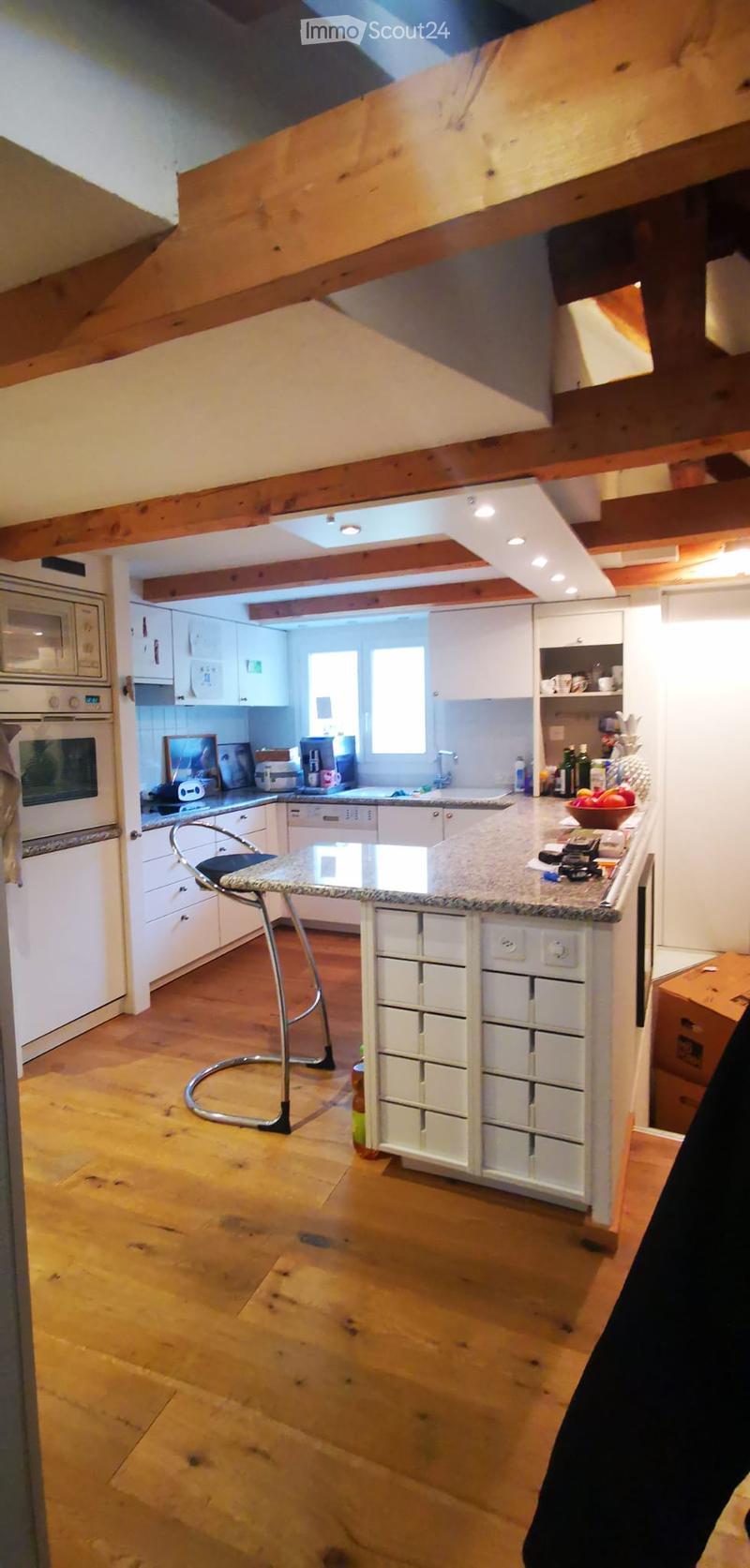 Modern kitchen with white cabinets, granite countertop, wooden floor, and stainless steel appliances including an oven, microwave, and dishwasher. A single stool is placed near the island.