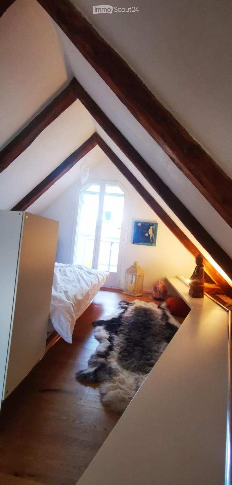 Cozy attic bedroom with wooden slanted ceilings, a bed, a fur rug, a small table with a candle, a decorative sculpture, and a painting on the wall.