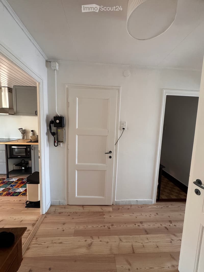 Empty room with wooden flooring, white walls, and a white door. The adjacent kitchen area is visible with cabinets, a microwave, and a countertop.