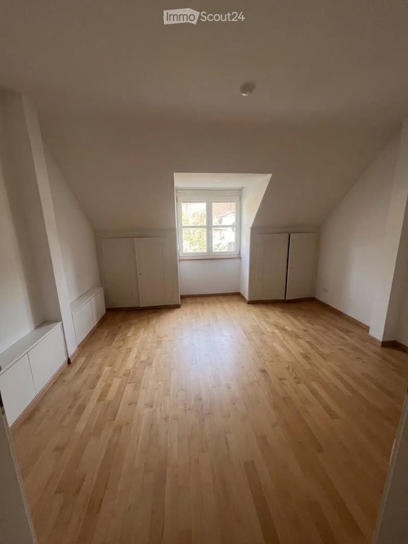 Empty attic room with wooden flooring, white walls, built-in cabinets, and a single window letting in natural light.