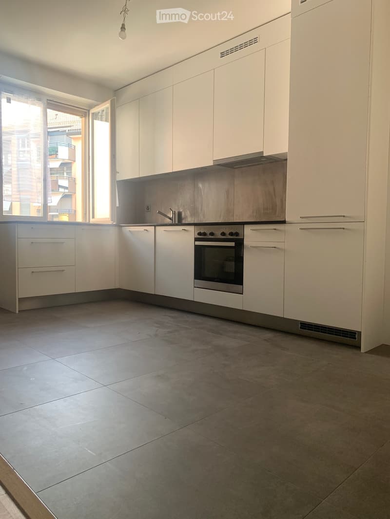 Modern kitchen with white cabinets, stainless steel oven, and tiled floor. Features a large window with a view of the street.