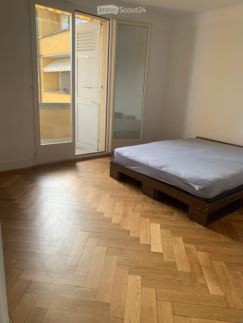 Bedroom with wooden herringbone flooring, a single bed with white bedding, large sliding glass doors leading to a balcony, and a mirror on the wall.