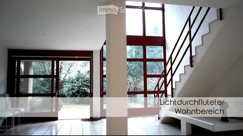 Bright, modern hallway with large windows and a staircase leading upwards. The space features white walls, tiled flooring, and a central column.
