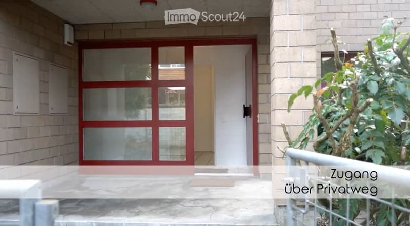 Entrance to a building with a red-framed glass door, white walls, and a small garden area with plants and a metal railing.