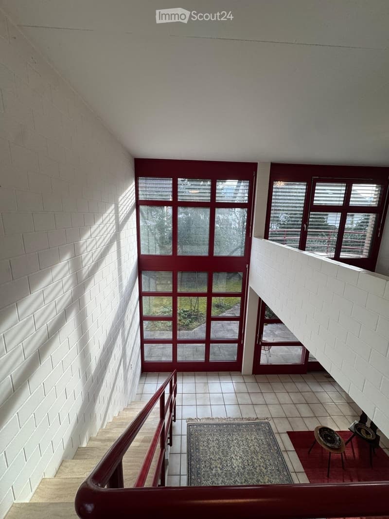 Modern hallway with white brick walls, tiled floor, large windows, and a small seating area with a stool and table.