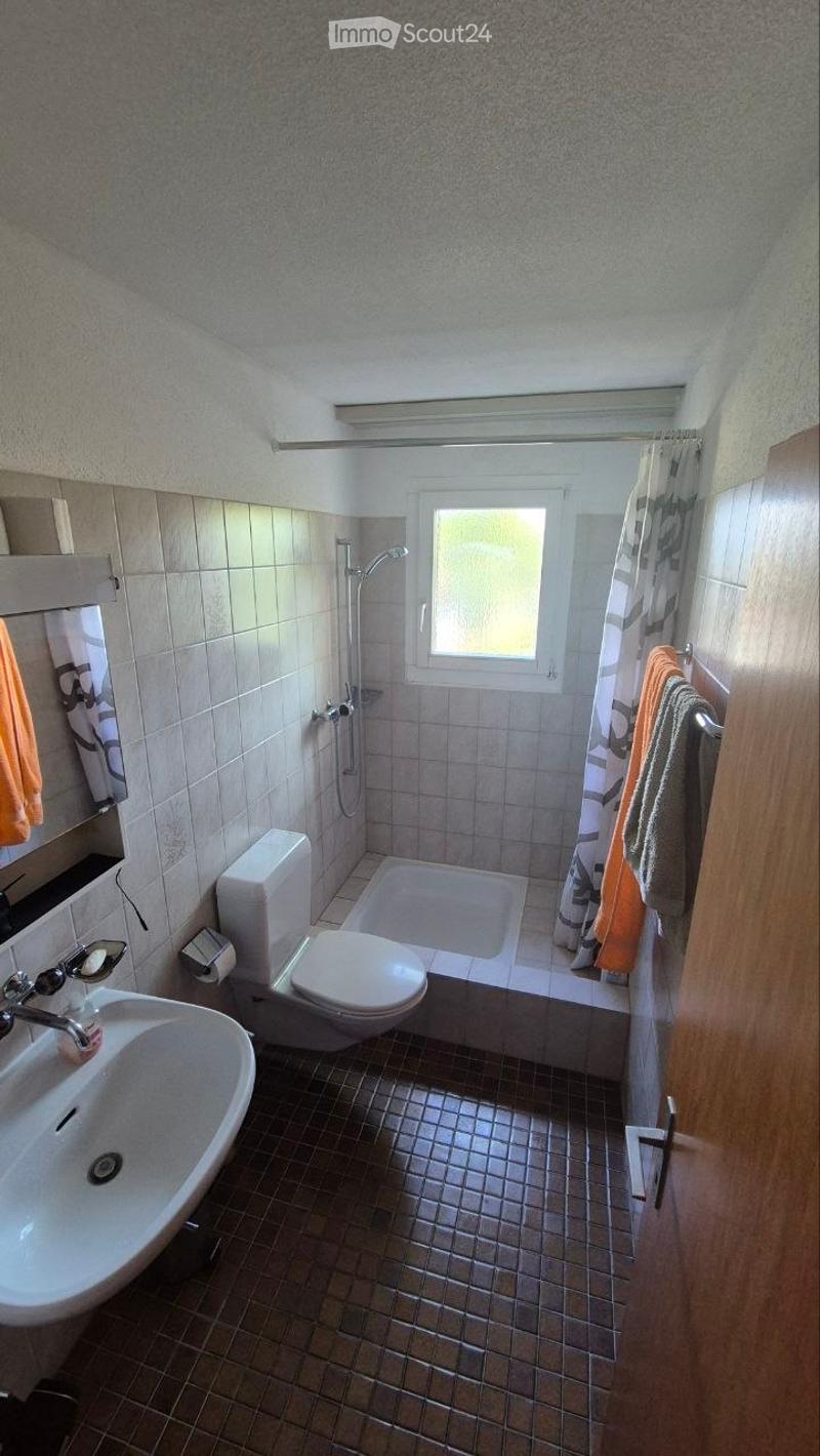 Bathroom with a sink, toilet, bathtub, shower, and towels hanging on the wall. The room has tiled walls and a window letting in natural light.