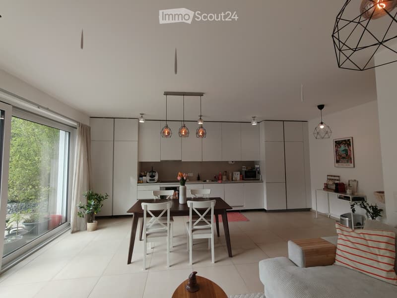 Modern open-plan living and dining area with white cabinets, pendant lights, a sofa, coffee table, dining table with chairs, and a kitchen area with appliances.