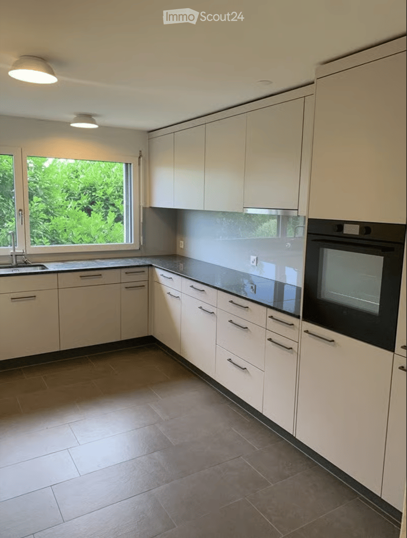 Modern kitchen with white cabinets, stainless steel appliances, and a large window overlooking greenery.