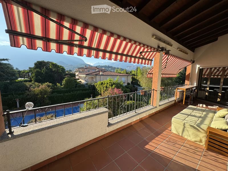 Balcony with tiled floor, covered by striped awnings, overlooking a scenic view of mountains and a swimming pool below.