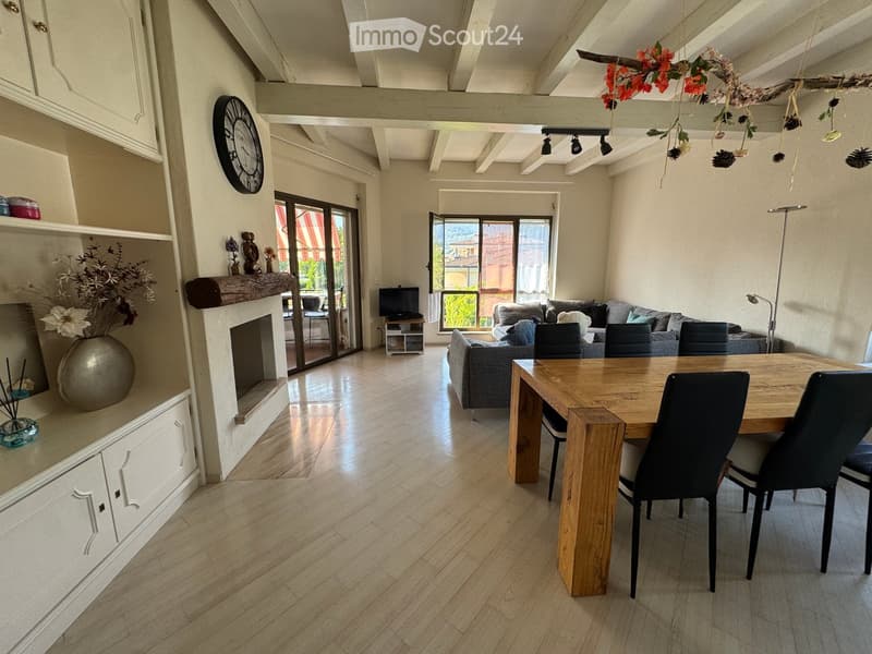 Open-plan living and dining area with wooden flooring, a fireplace, a sofa, a dining table with chairs, and a television. Large windows offer a view of the outdoor area.
