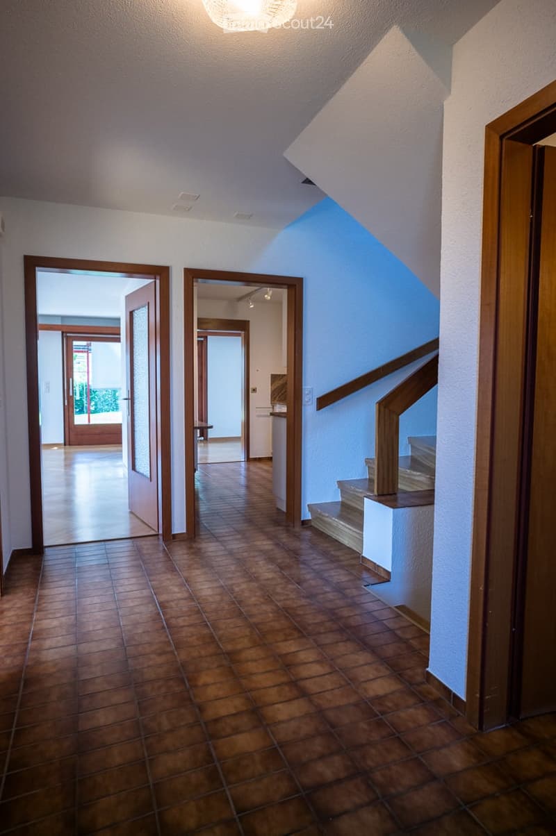 Spacious hallway with brown tiled flooring, white walls, and wooden doors leading to other rooms. A staircase with wooden railings ascends to an upper level.