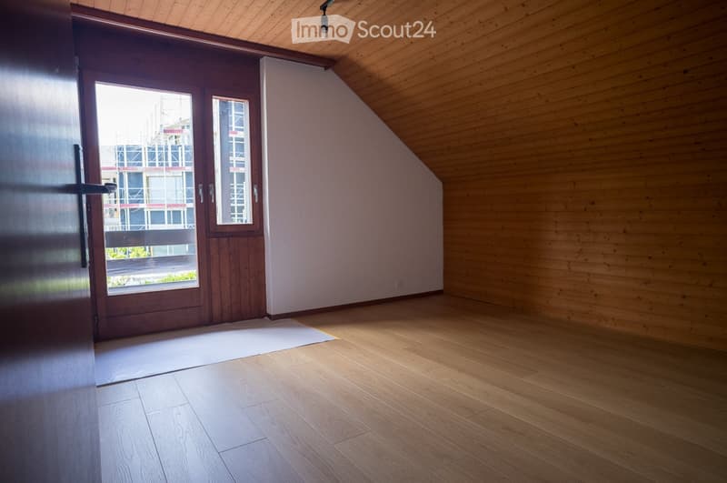 Empty attic room with wooden walls and floor, large windowed door leading to a balcony, and a small white rug on the floor.