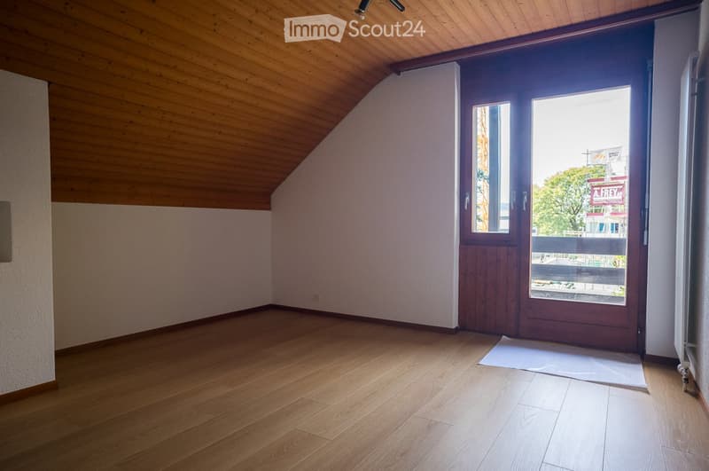 Empty room with wooden flooring and slanted wooden ceiling, featuring a large windowed door leading to a balcony.