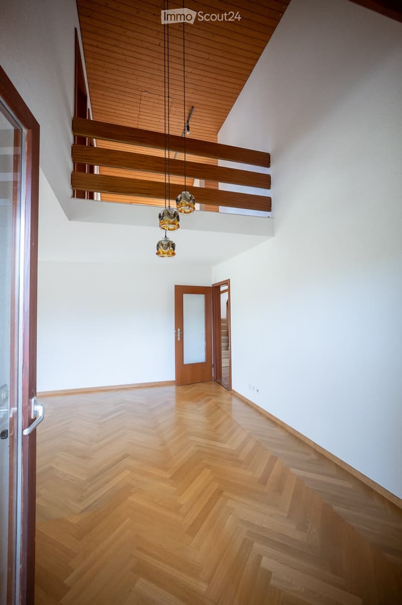 Empty hallway with wooden herringbone flooring, white walls, and a wooden ceiling with exposed beams. Features pendant lights and a wooden door leading to another room.