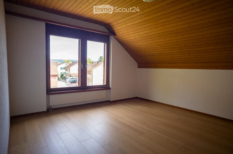 Empty attic room with wooden flooring, slanted wooden ceiling, and large window offering a view of the street and neighboring houses.