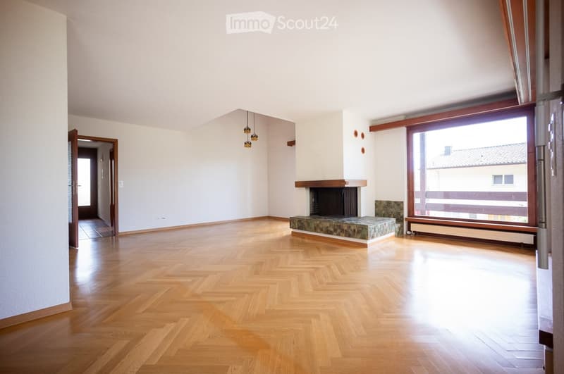 Empty room with wooden herringbone flooring, white walls, fireplace, large window, and pendant lights.