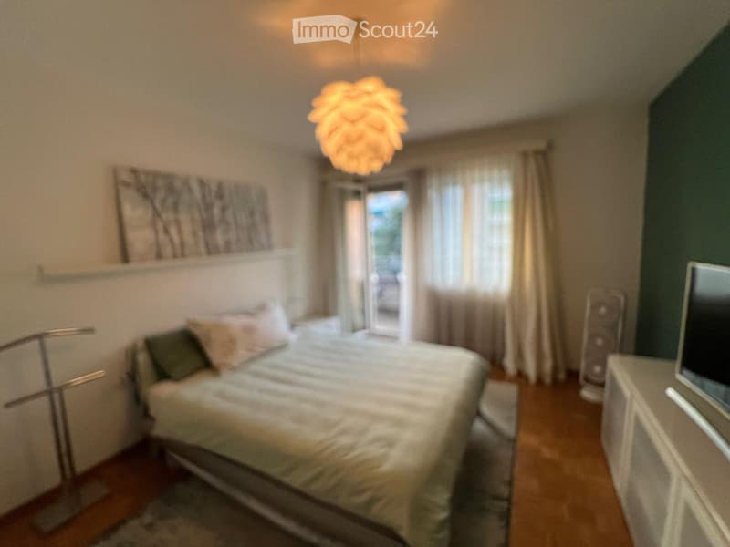 A well-lit bedroom with a large bed, nightstand, and a hanging lamp. The room features wooden flooring, a large window with curtains, and a decorative wall piece above the bed.