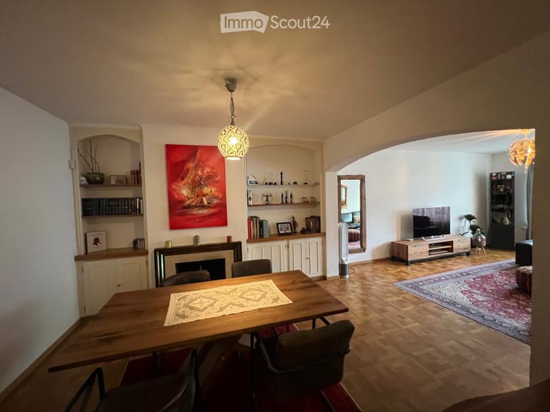 A well-furnished living and dining area with a wooden dining table, chairs, shelves, a fireplace, a large mirror, a television, and a fan. The space features a mix of modern and traditional decor, including a colorful painting and a patterned rug.