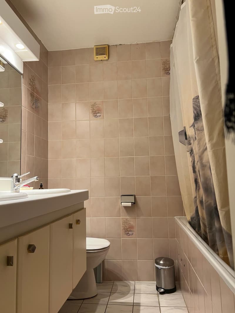 Bathroom with beige tiled walls and floor, double sink, toilet, and a shower curtain partially visible.