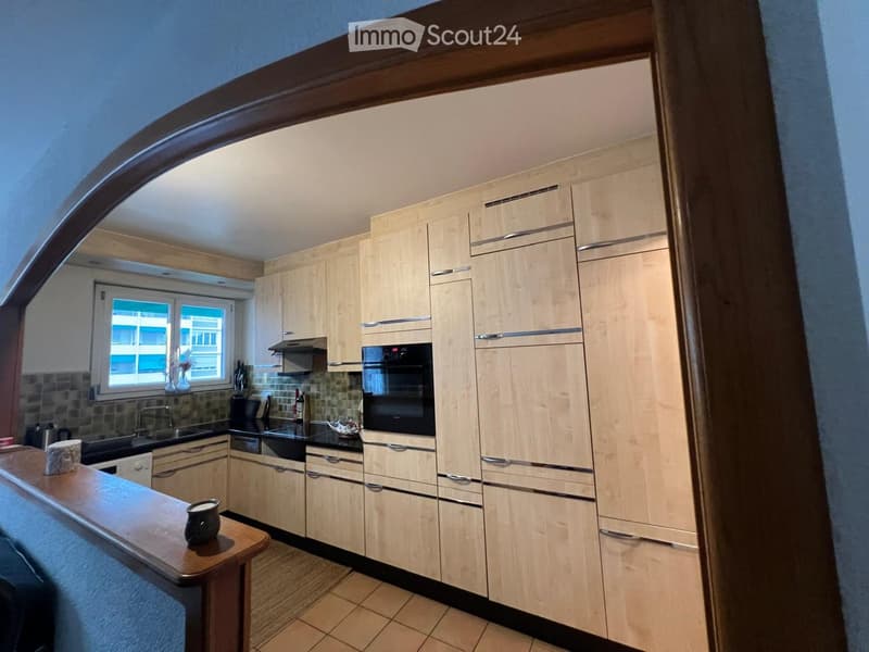 Modern kitchen with light wood cabinets, stainless steel appliances, and a black countertop. Features include an oven, sink, and various small items on the counter.