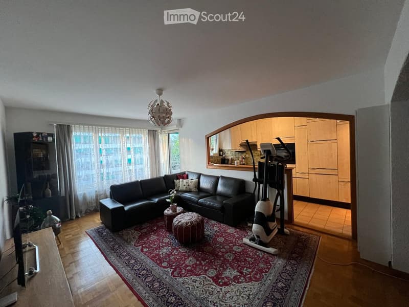A modern living room with a black leather sectional sofa, two ottomans, a round stool, a patterned rug, an exercise bike, a large window with sheer curtains, and a kitchen area visible in the background.