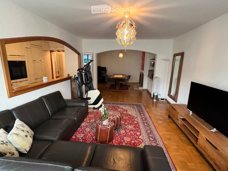 A modern living room with a black leather sofa, ottoman, coffee table, screen, mirror, fan, exercise bike, and a dining area with a table and chairs.