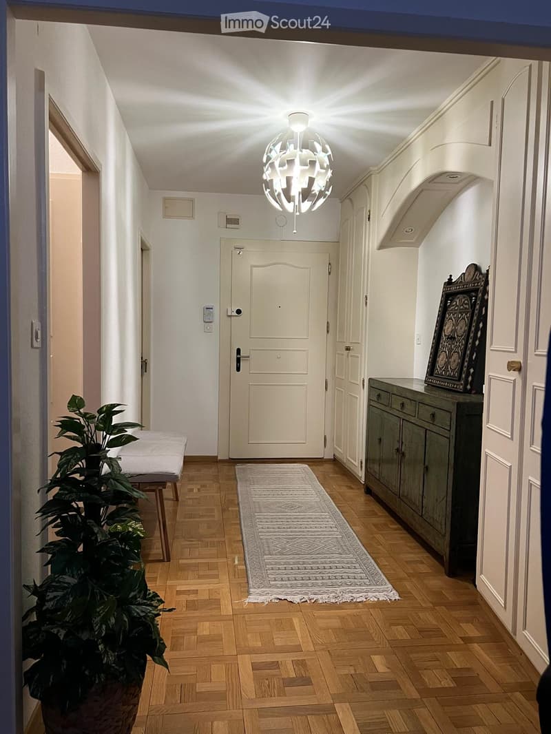 Hallway with wooden flooring, white walls, a modern chandelier, a small bench, a potted plant, a rug, and a cabinet with a framed artwork on top.