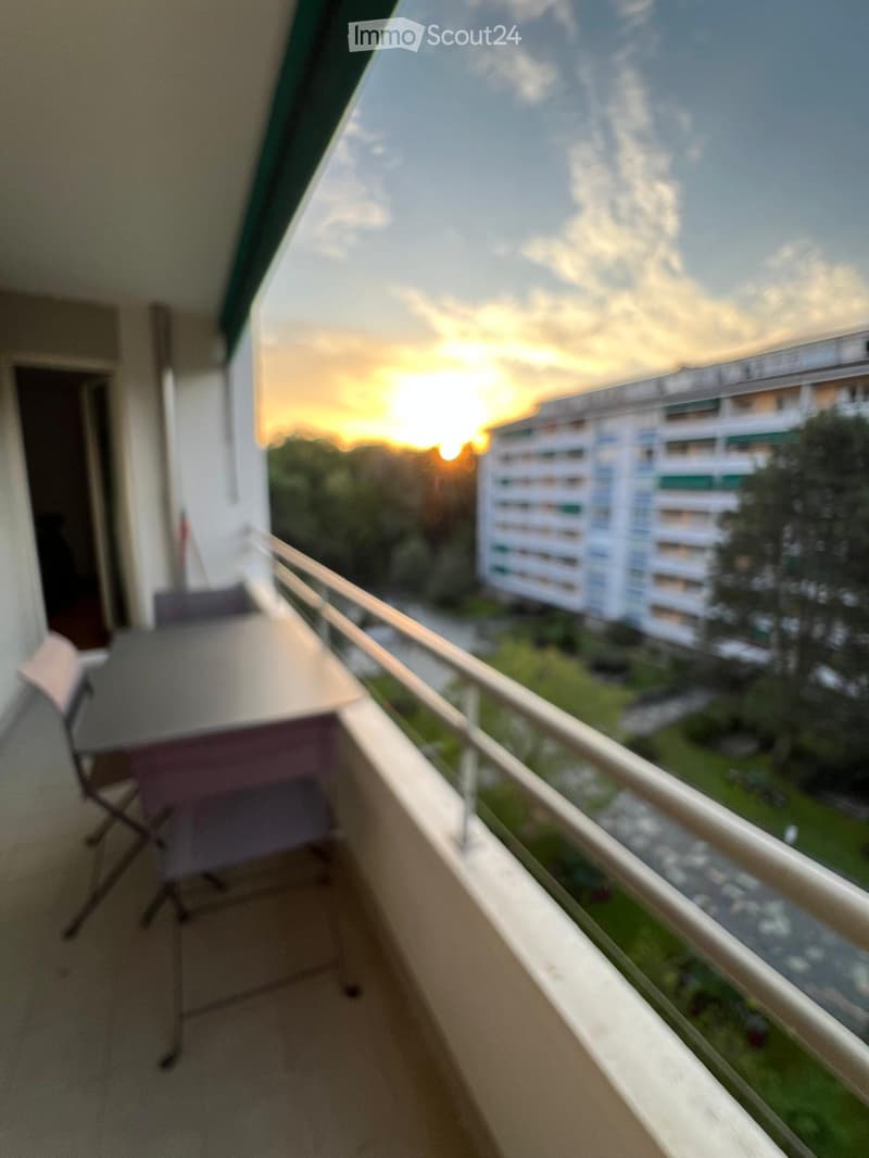 Balcony with a metal railing, a table, and a chair, overlooking a cityscape at sunset.