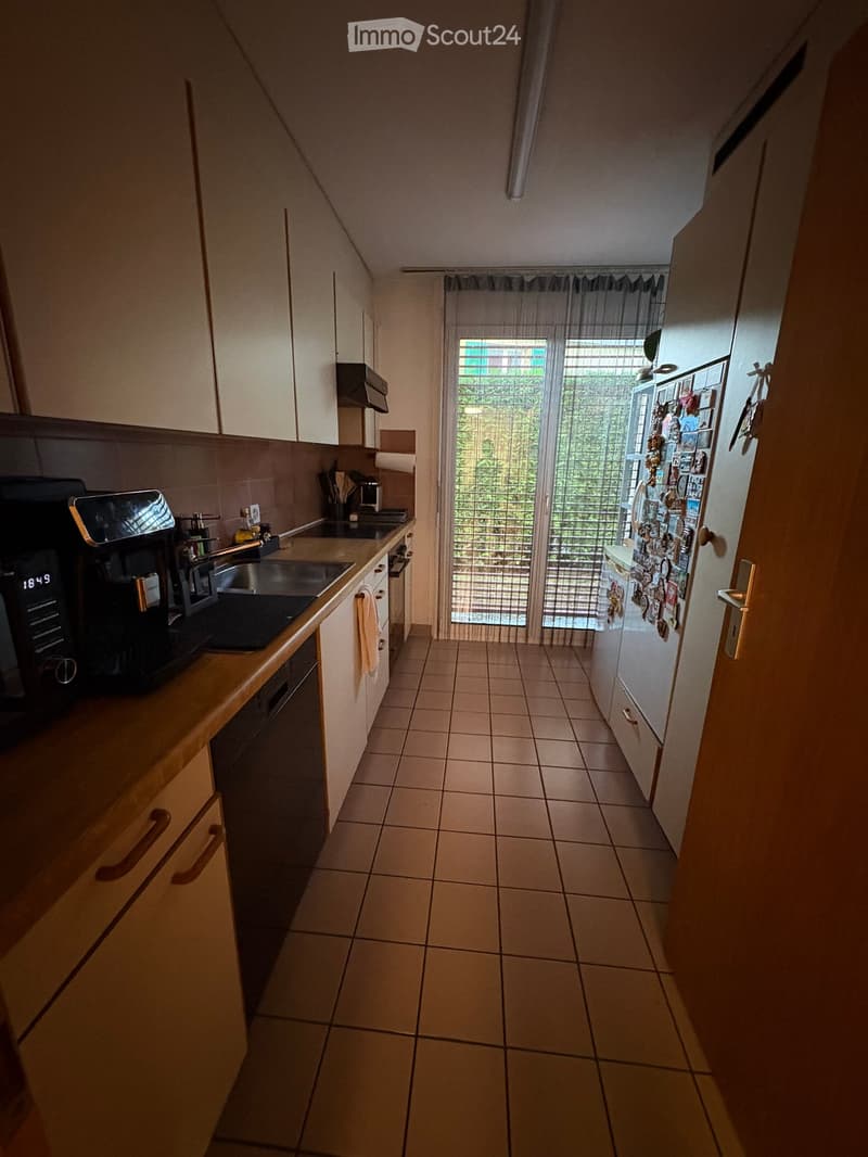 Modern kitchen with tiled floor, white cabinets, stainless steel appliances, and a view of greenery through a glass door.