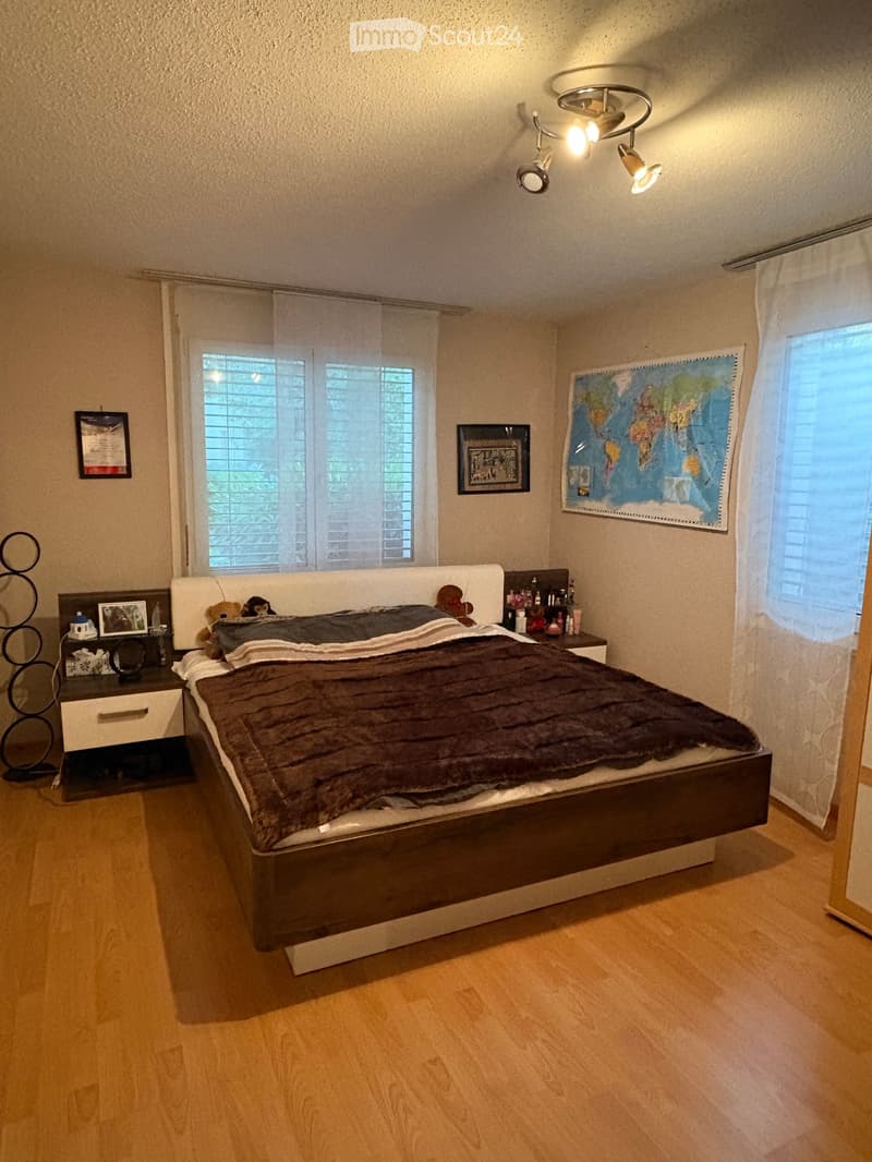 A well-lit bedroom with a large bed, nightstand, mirror, framed picture, and a world map on the wall. The room features wooden flooring and large windows with blinds.