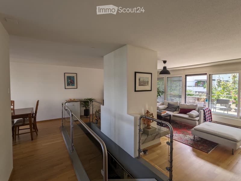 Open-plan living and dining area with wooden flooring, large windows, a sofa, ottoman, dining table, chairs, candle holder, picture frames, and a plant.