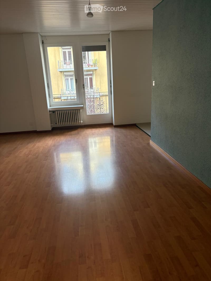 Empty room with wooden flooring, white walls, and a large window with a view of a balcony and building exterior.